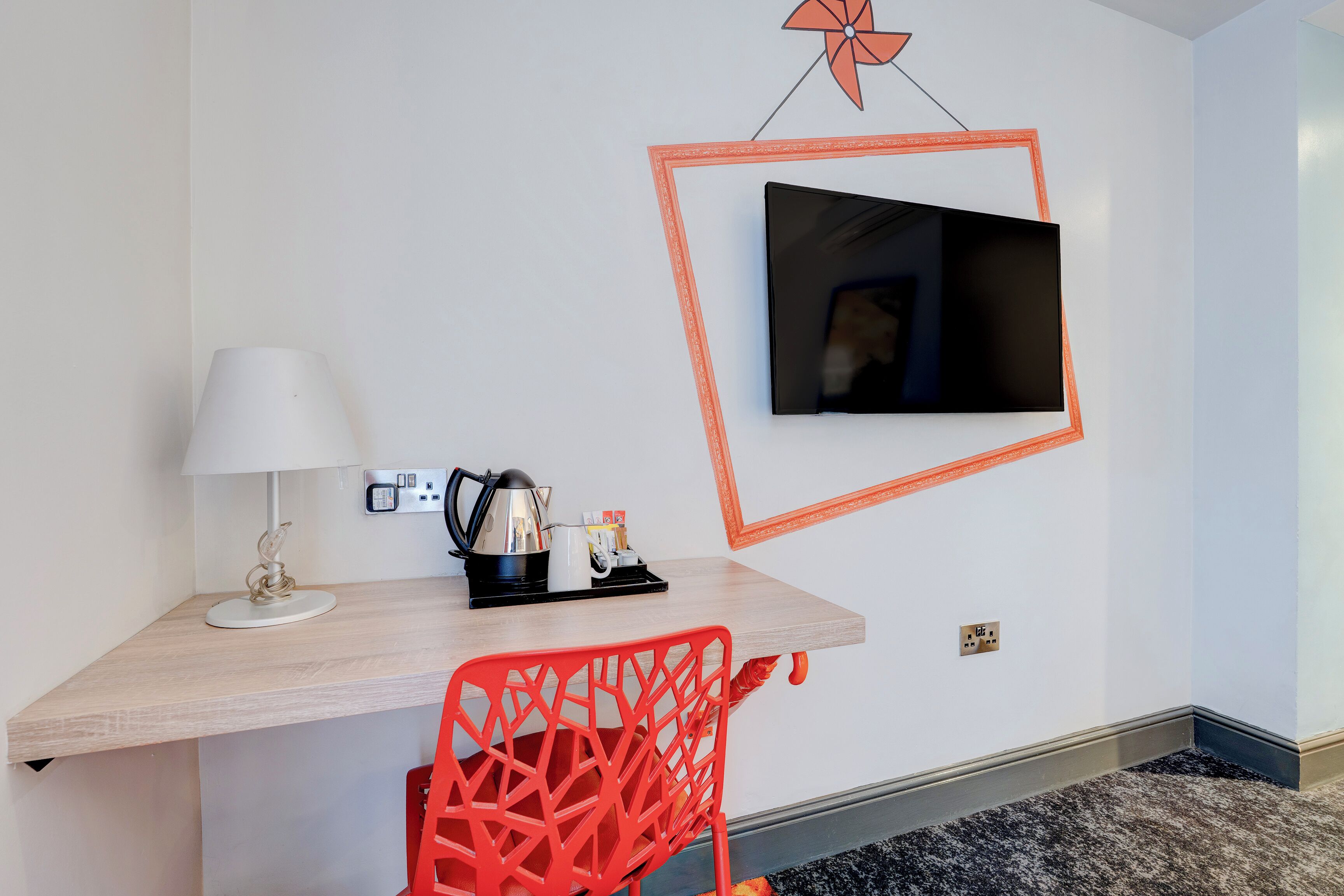 A hotel room with a wooden desk, a red chair, a white lamp, a television mounted on the wall, and a small coffee station.