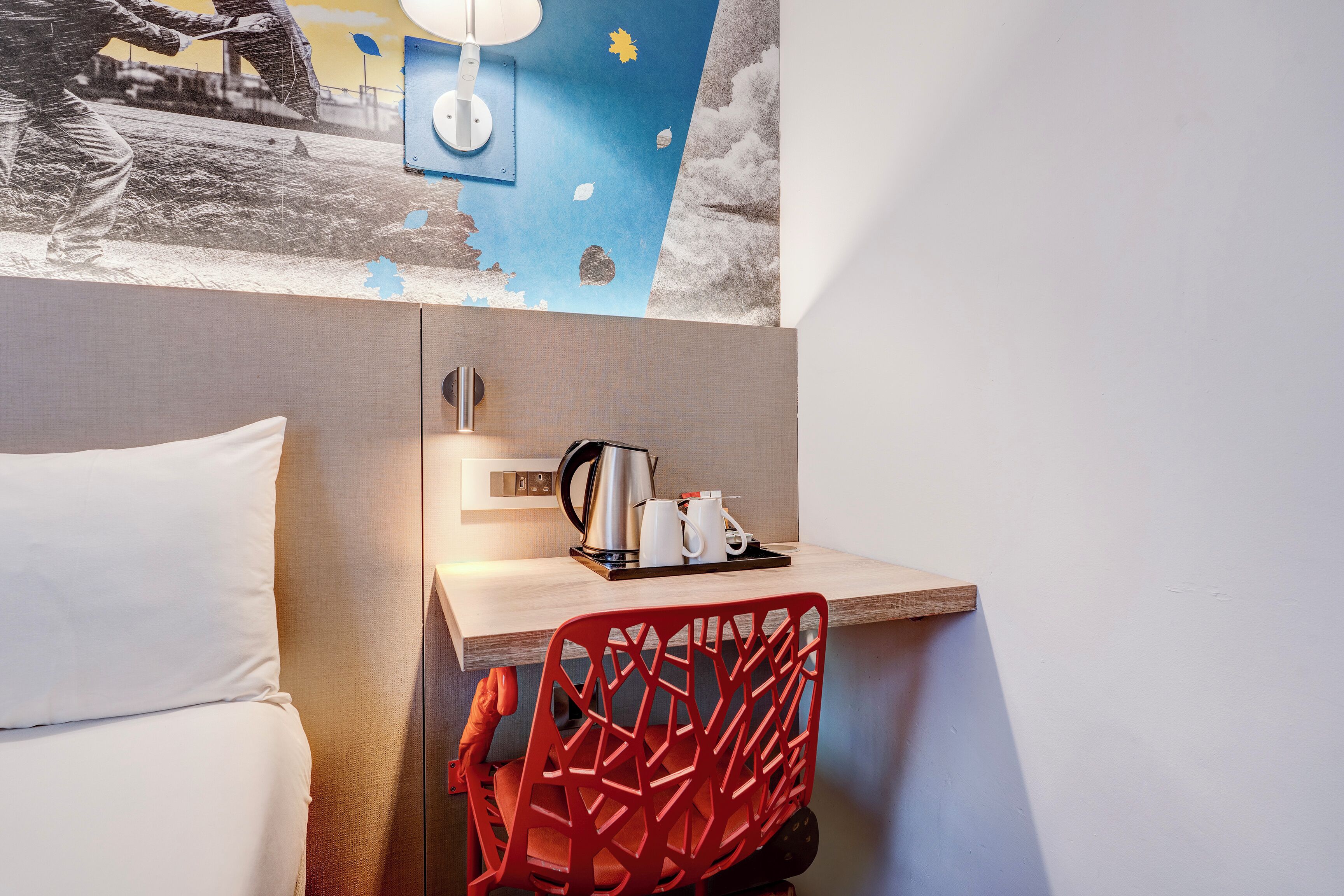 Habitación de hotel con cama, escritorio con tetera y tazas, una silla roja y una pared con un mural.