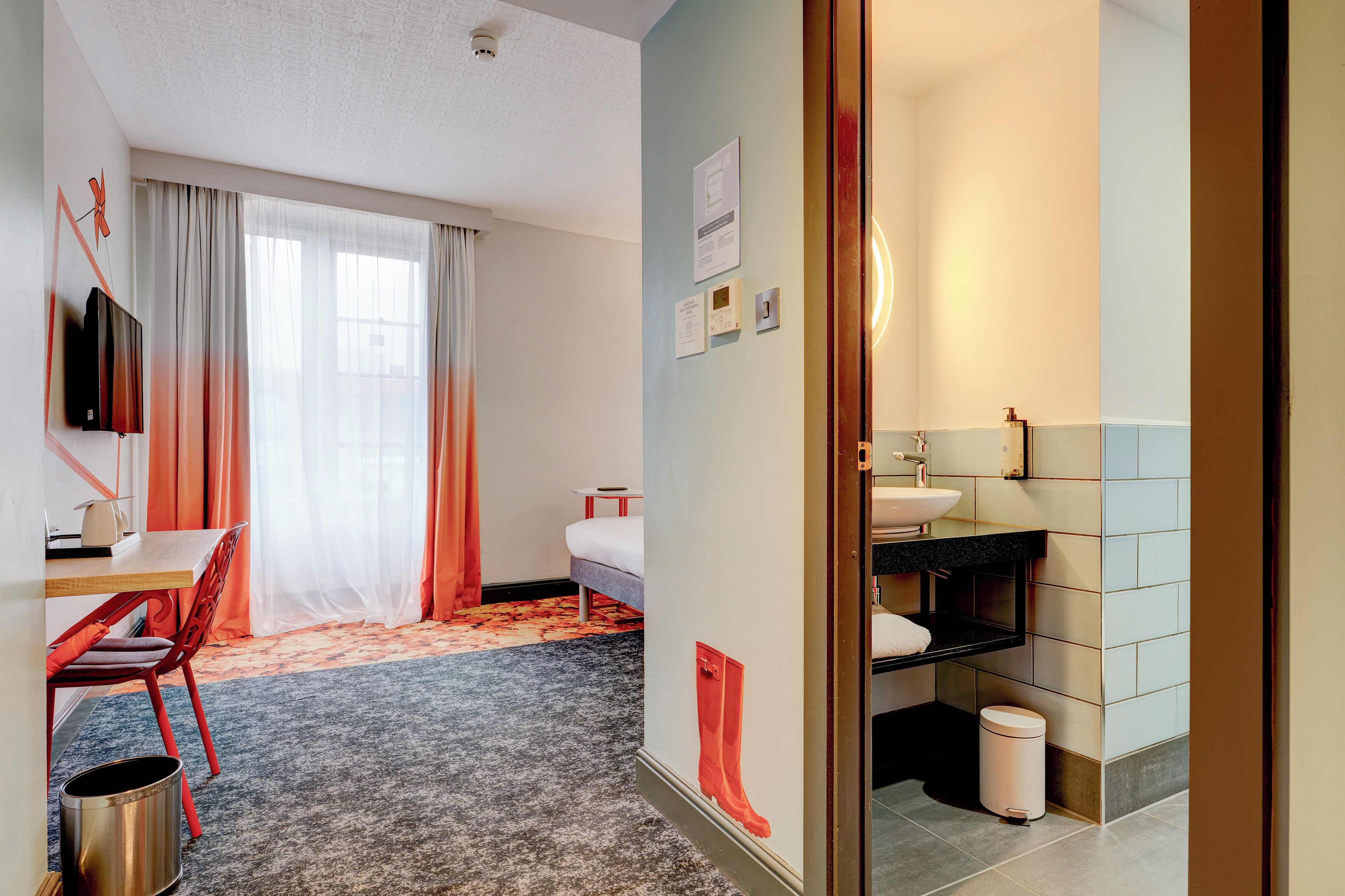 A hotel room with a bed, a desk with a chair, a TV, a bathroom with a sink and toilet, and a trash bin.