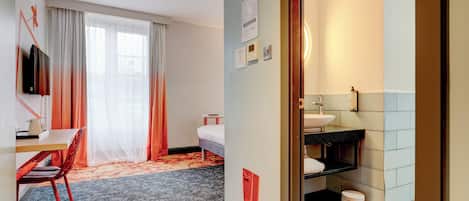 A hotel room with a bed, a desk with a chair, a TV, a bathroom with a sink and toilet, and a trash bin.