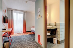 Habitación de hotel con cama, escritorio con silla, televisor, baño con lavamanos e inodoro, y un cubo de basura.