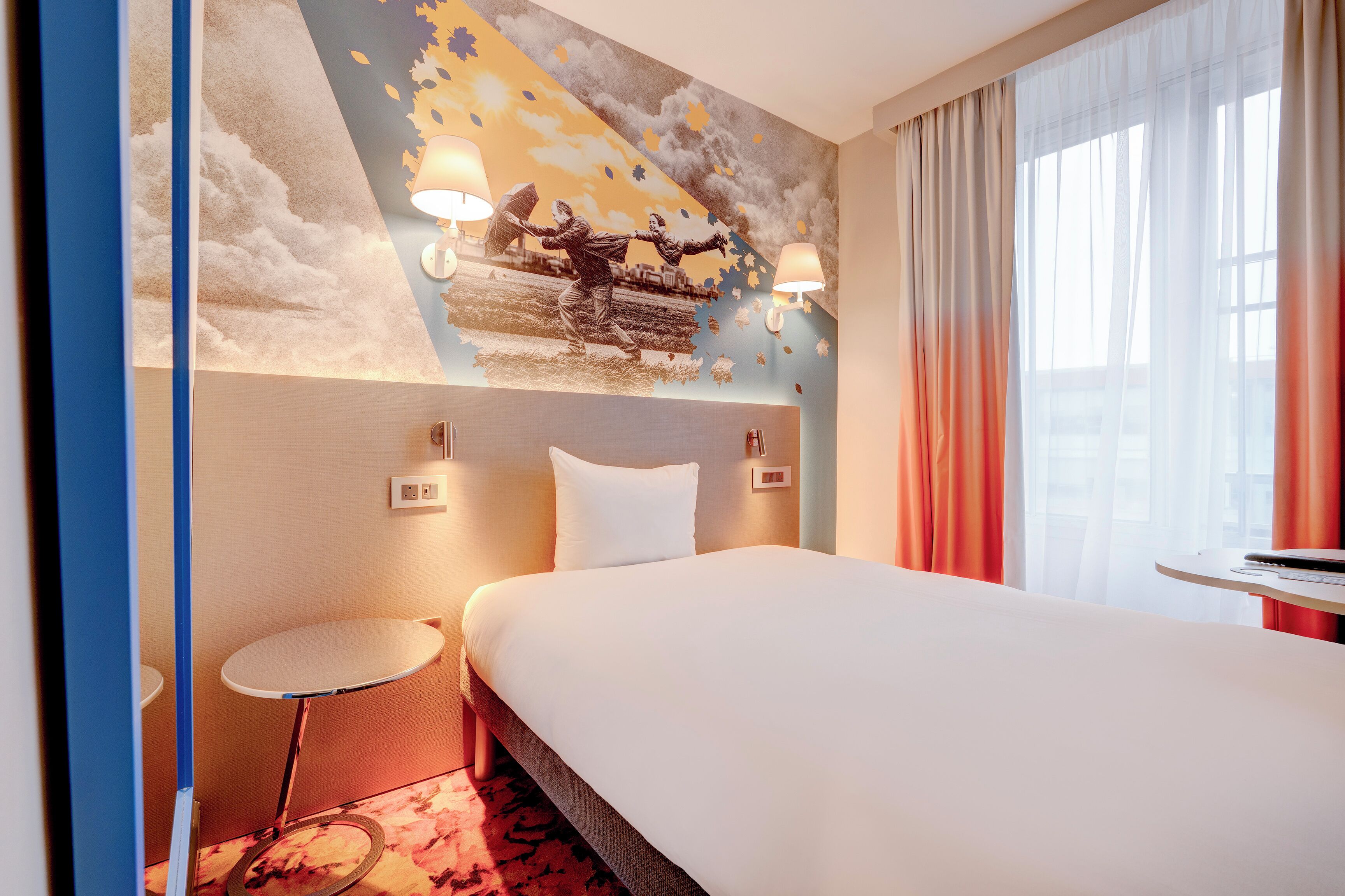 A hotel room with a large bed, a small round table, a wall-mounted lamp, and a mural of a train scene.