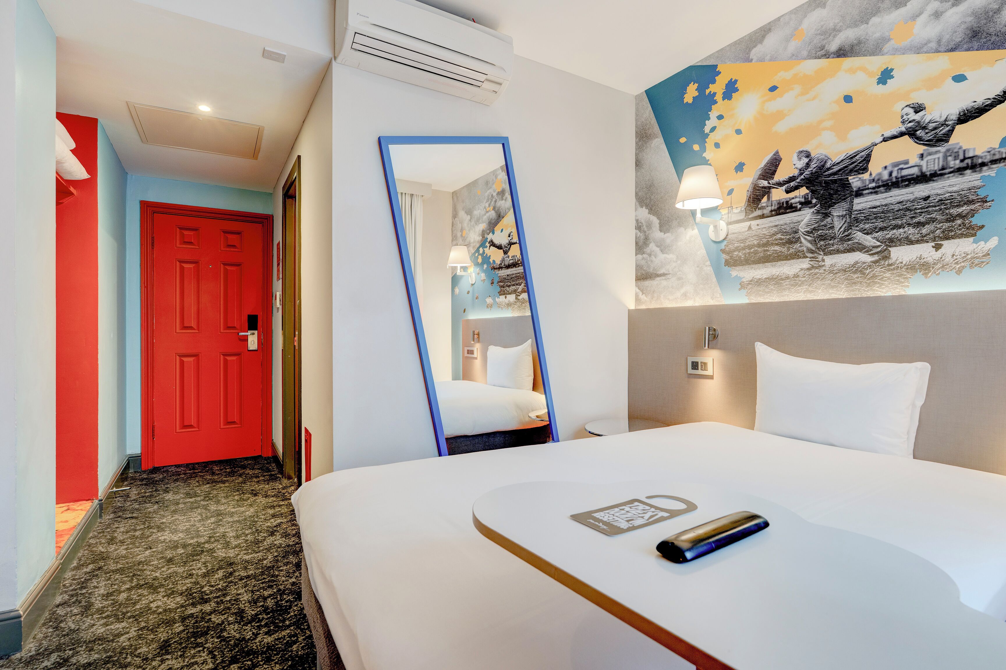 Ein Hotelzimmer mit einem Bett, einem Spiegel, einem Gemälde und einer roten Tür.
