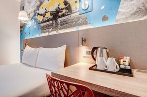 Habitación de hotel con cama, una mesa de madera con bandeja de tazas de café y una tetera, y un mural en la pared.
