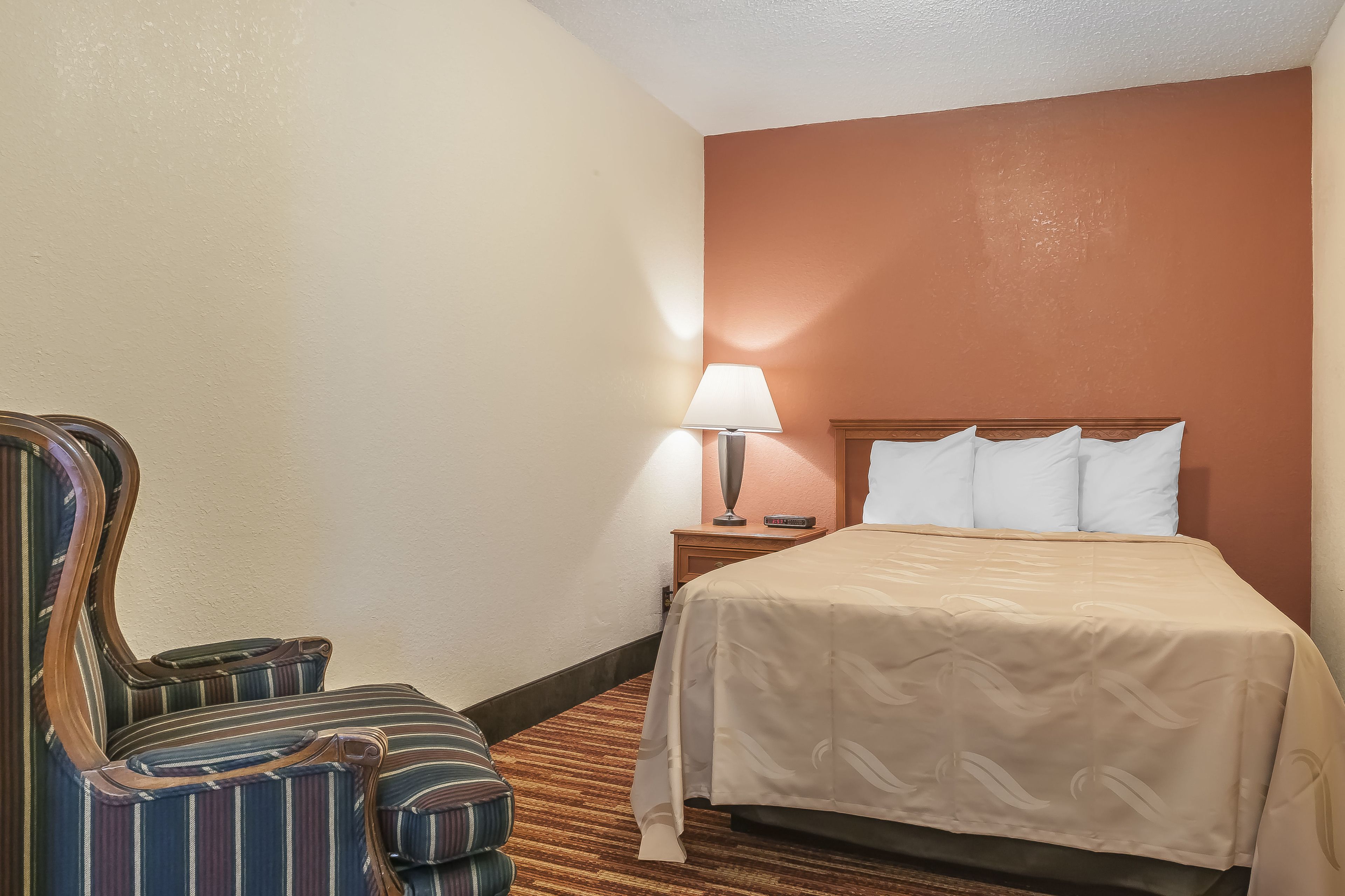 A hotel room with a bed, a nightstand with a lamp, and a striped armchair.