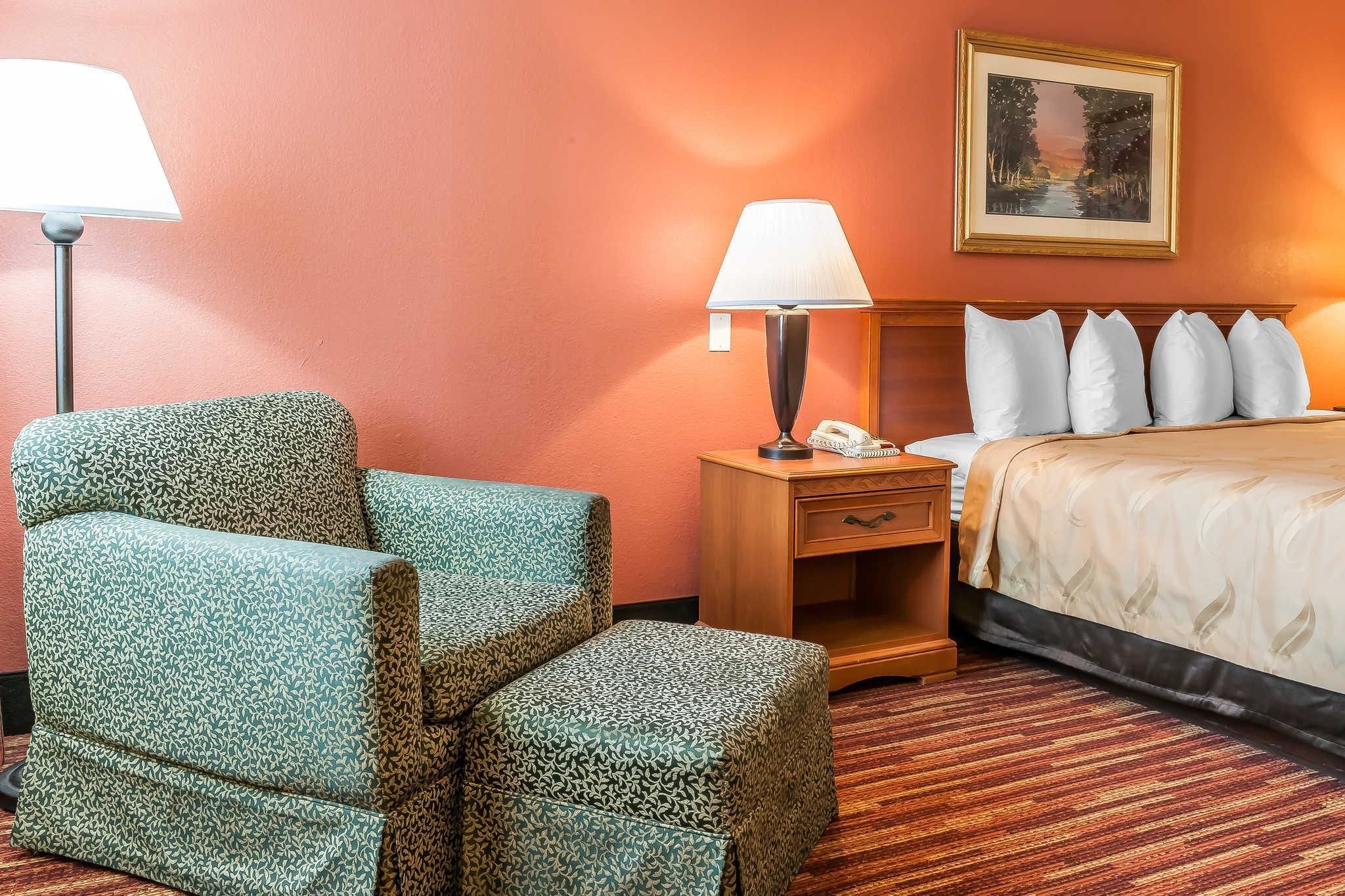 Habitación de hotel con un sillón estampado, una mesita de noche con lámpara, una cama con almohadas blancas y un cuadro en la pared.
