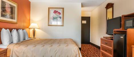 A hotel room with a bed, a desk with a lamp, a dresser, a television, and a framed picture on the wall.