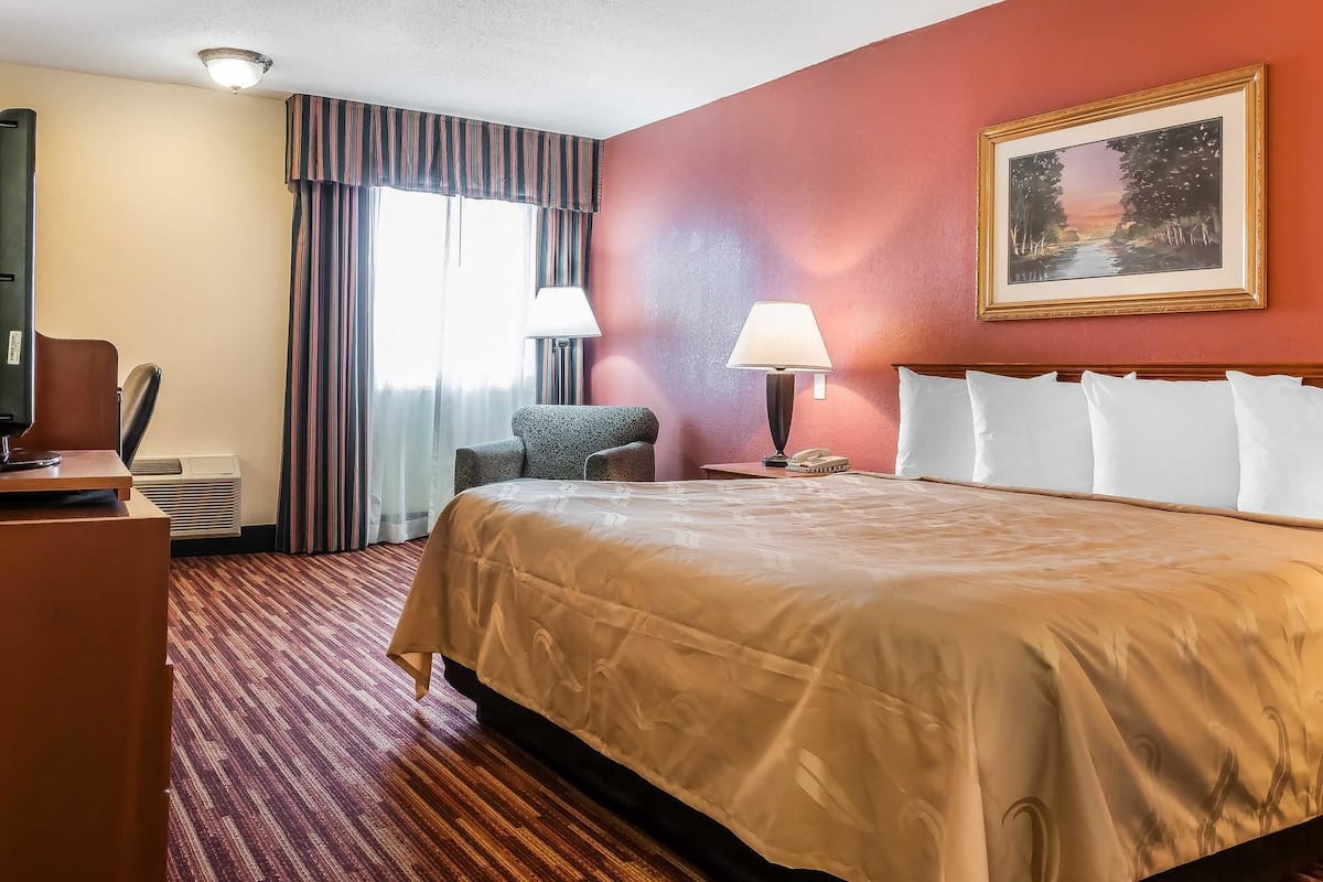 A hotel room with a large bed, a desk, a chair, a lamp, and a painting on the wall.