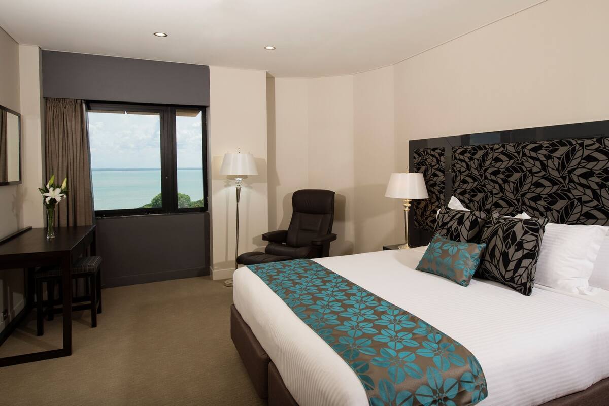 A hotel room with a bed, a desk, a chair, a window with a view, and a patterned headboard.