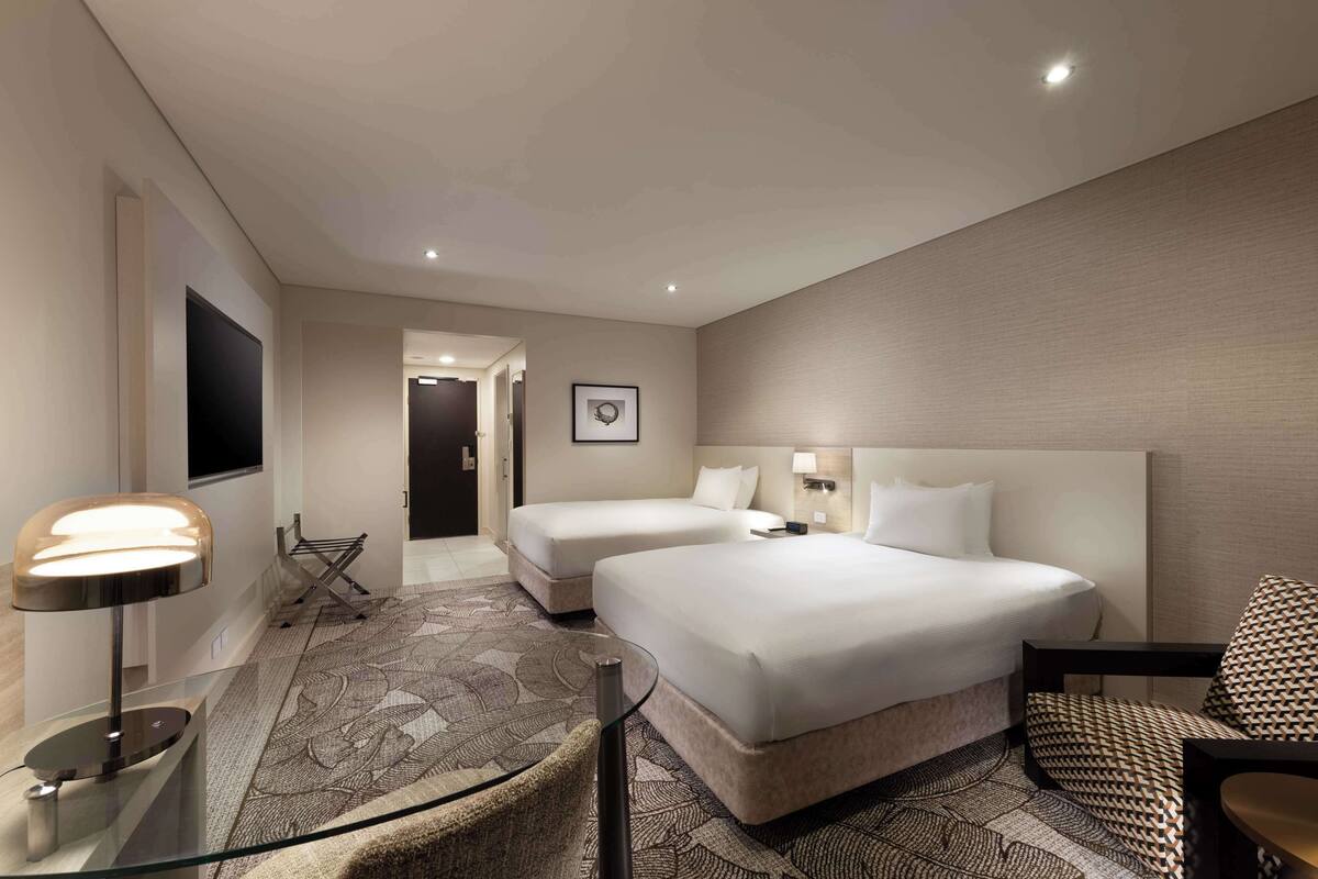 A hotel room with two beds, a glass table, a lamp, a patterned rug, and a chair.