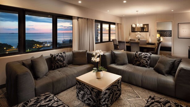 Un salon moderne avec un grand canapé d’angle, une table basse et une vue sur la ville au coucher du soleil.