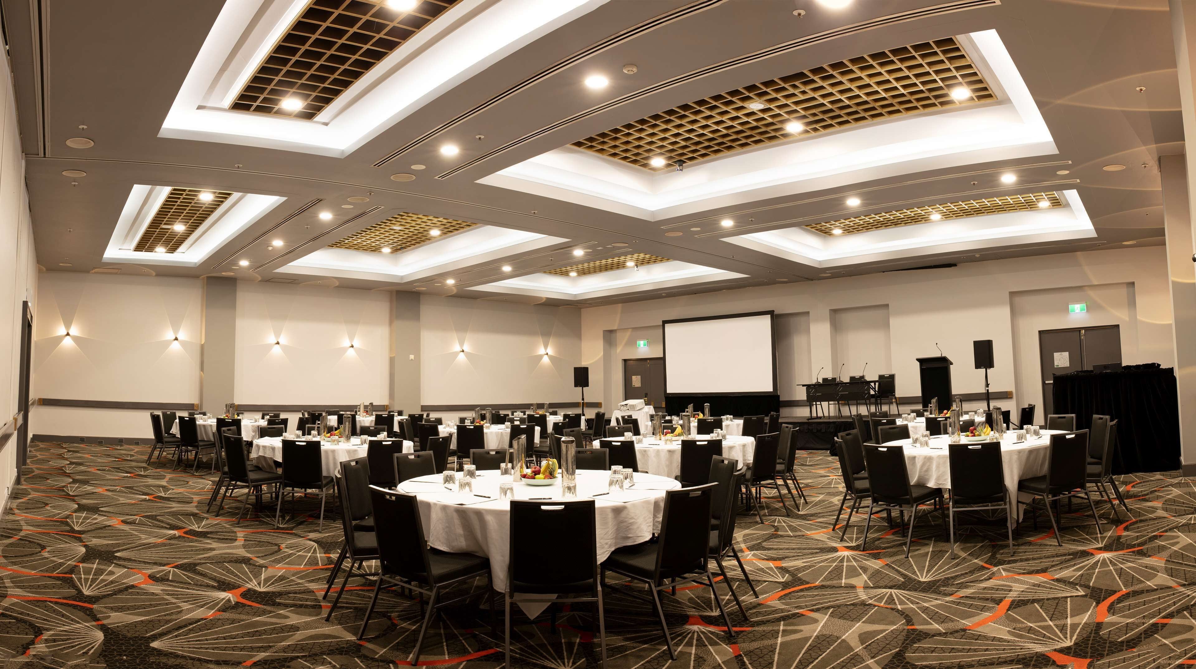 A conference room with multiple tables and chairs, a projector screen, and a stage with a podium.