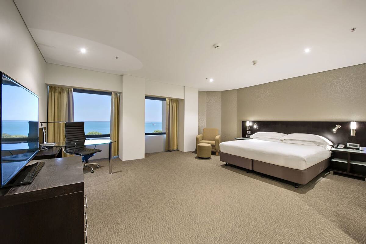 A hotel room with a large bed, a desk with a computer, a chair, and a view of the sea.