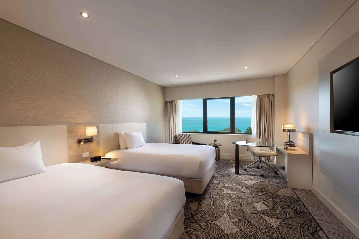 A hotel room with two beds, a desk, a chair, a TV, and a view of the ocean.