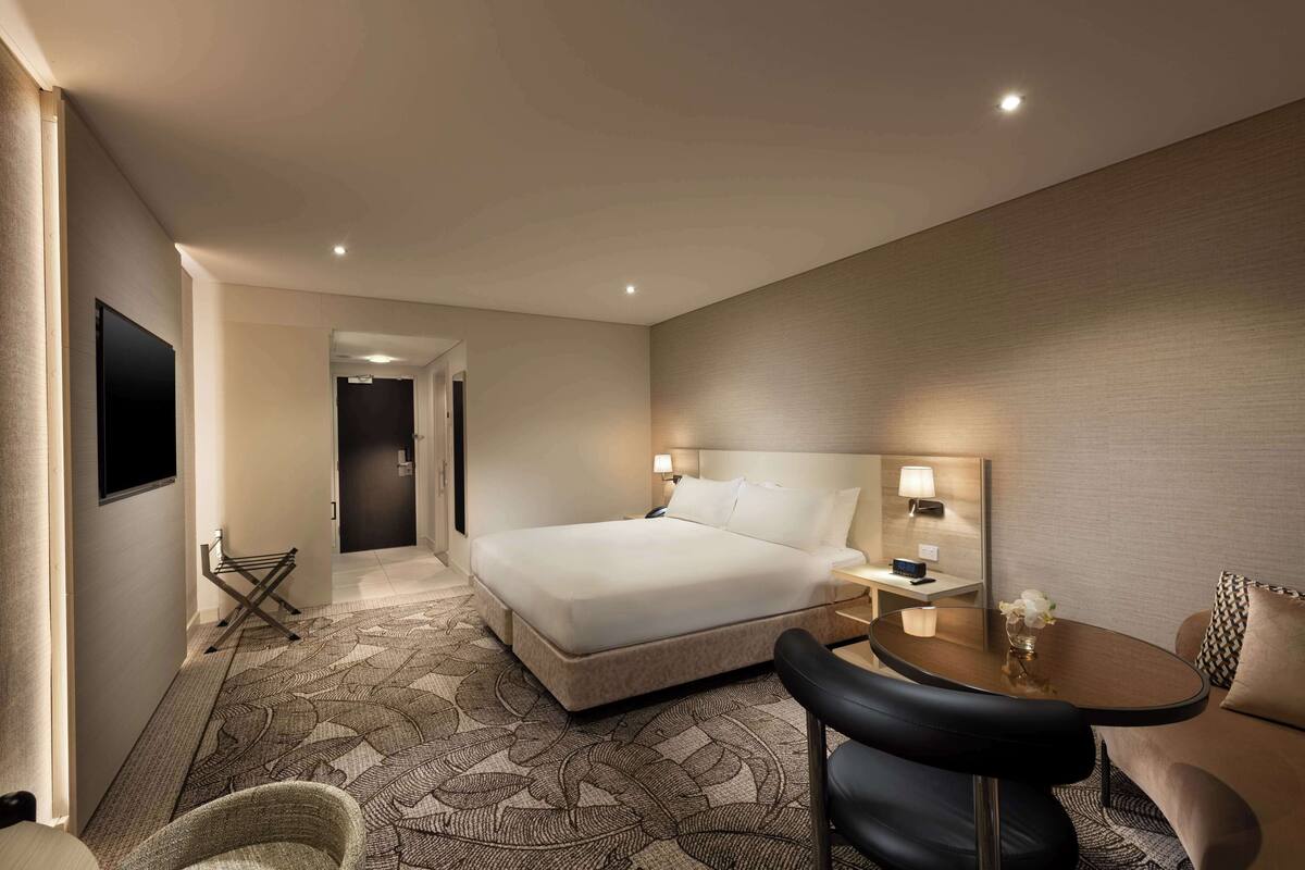 A hotel room with a large bed, a desk with a lamp, a chair, and a small table with a vase.