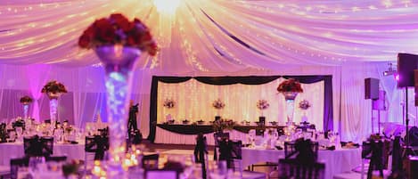 A banquet hall with tables set for a formal event, draped with white fabric and illuminated by string lights.