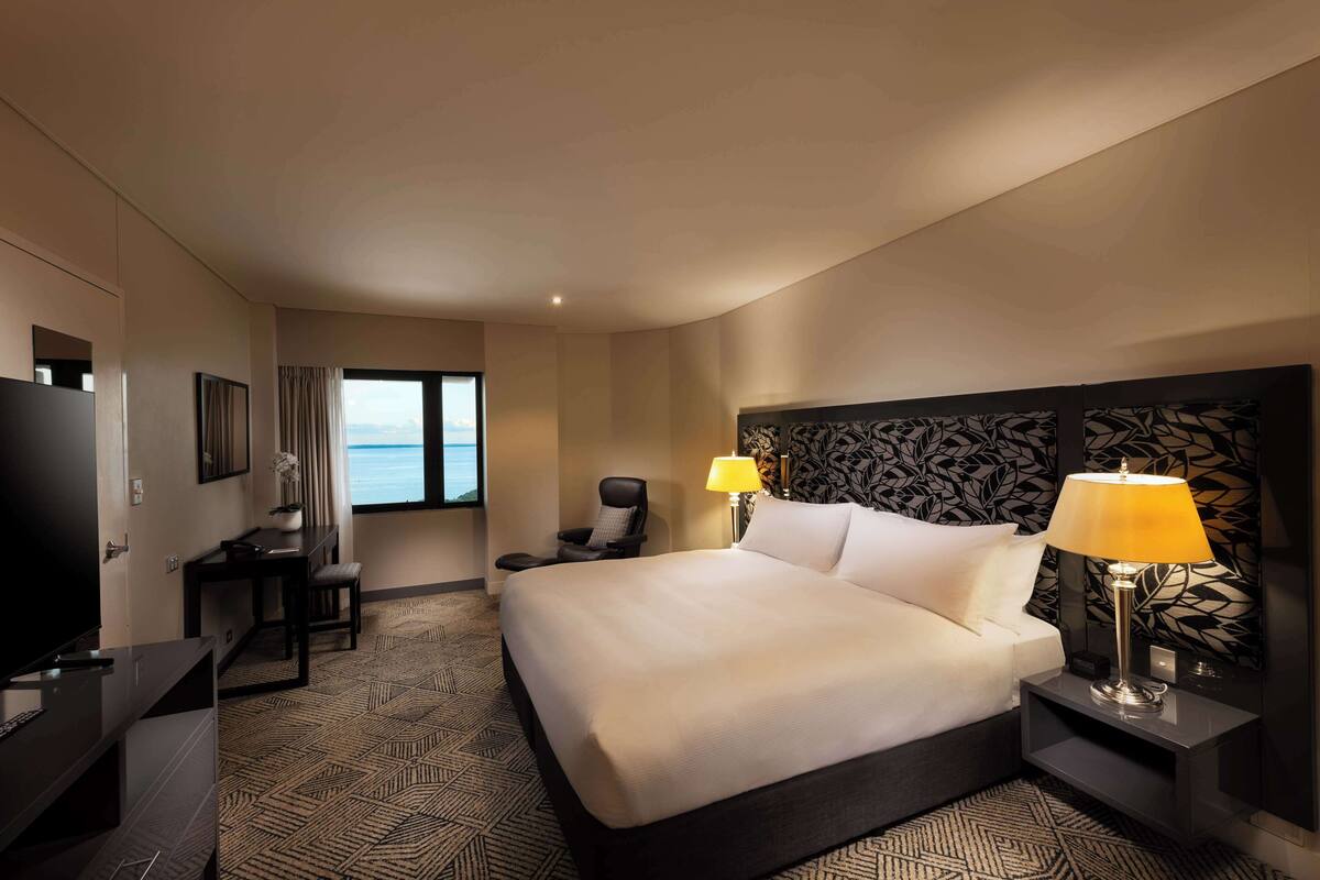 A hotel room with a large bed, a desk with a chair, a television, and a view of the sea through the window.
