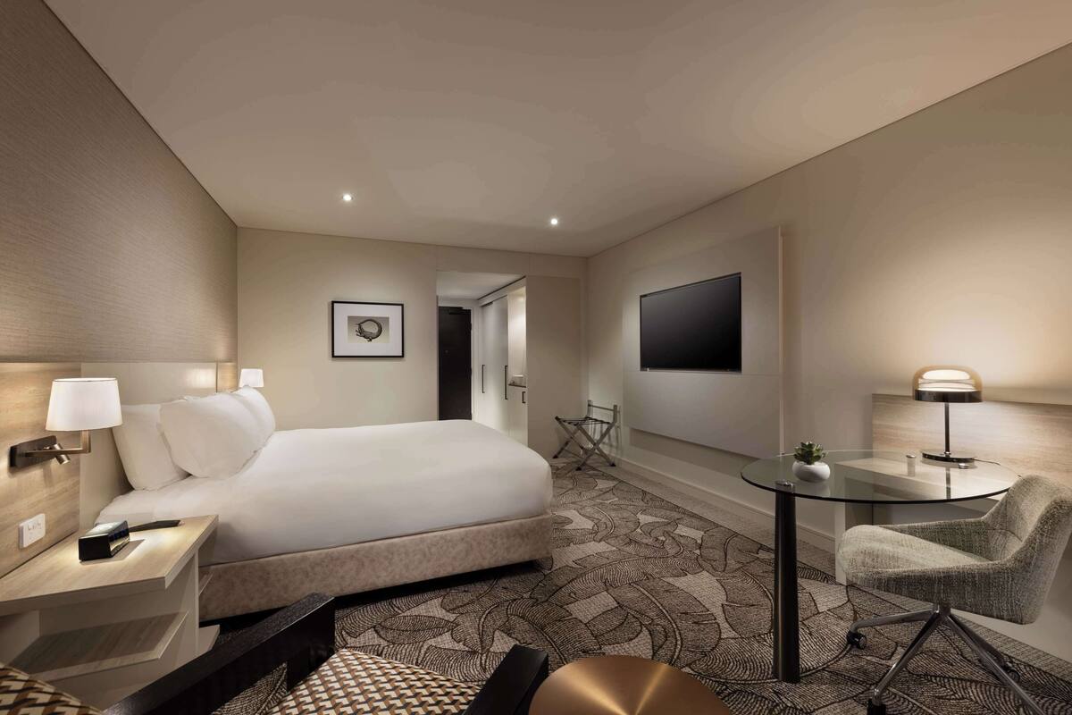 A hotel room with a bed, bedside table, lamp, chair, and a small table with a plant.