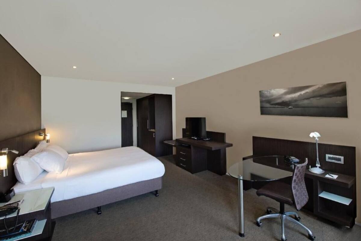 A hotel room with a bed, desk, chair, and a painting on the wall.