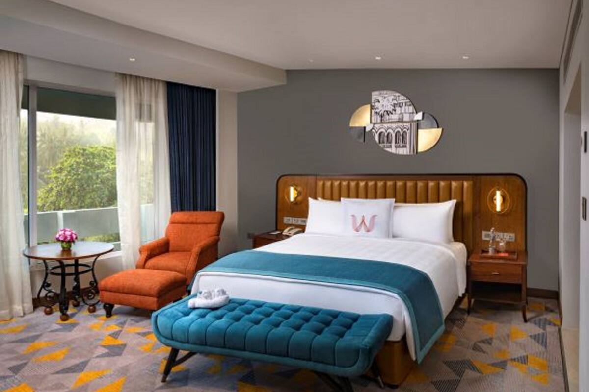 Une chambre d’hôtel avec un grand lit, une tête de lit en bois, une table de chevet avec une lampe, une petite table ronde ornée d’un vase de fleurs et une fenêtre avec des rideaux.