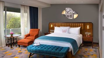 A hotel room with a large bed, a wooden headboard, a bedside table with a lamp, a small round table with a vase of flowers, and a window with curtains.