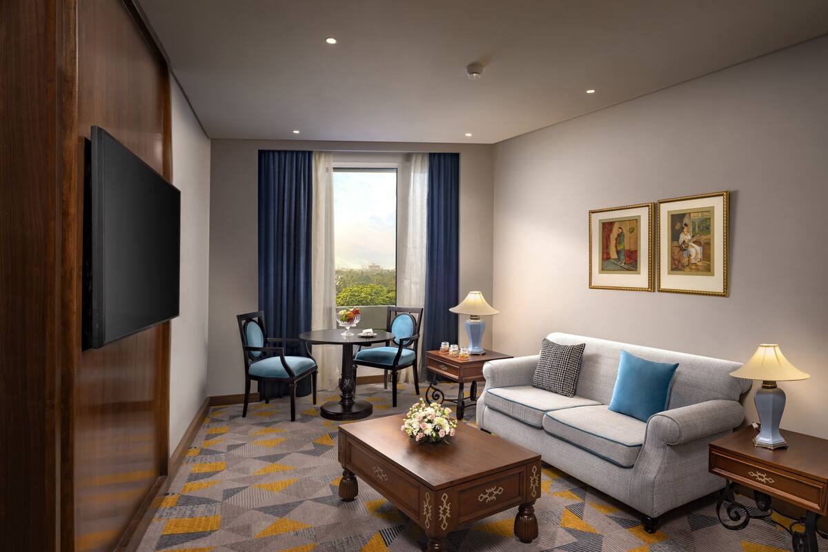 Une chambre d’hôtel équipée d’un téléviseur à écran plat, d’un canapé, d’une table basse agrémentée d’un arrangement floral, d’une table à manger avec des chaises et d’une grande fenêtre avec des rideaux.