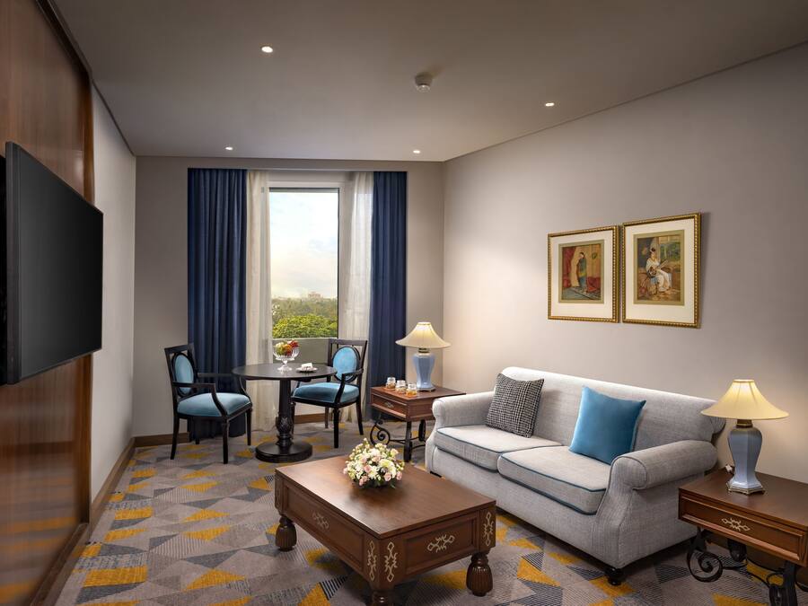 Quarto de hotel com televisão de tela plana, sofá, mesa de centro com arranjo floral, mesa de jantar com cadeiras e uma grande janela com cortinas.