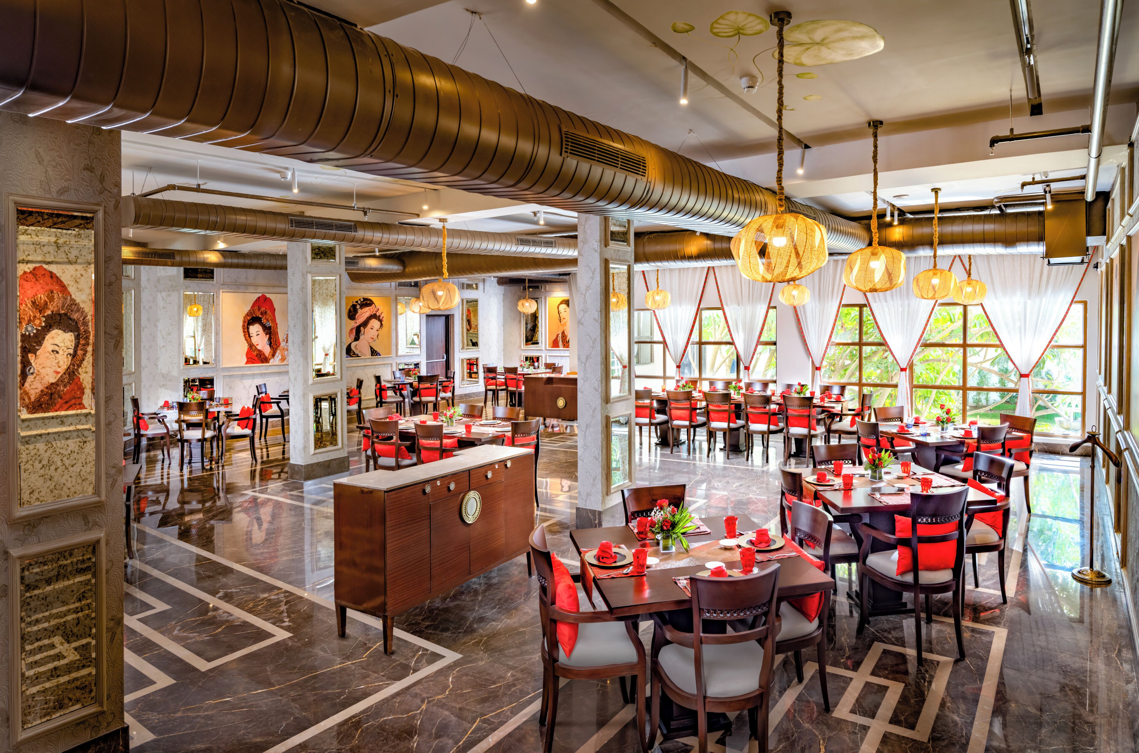A spacious hotel dining area with multiple tables and chairs, a central reception desk, and large windows allowing natural light.
