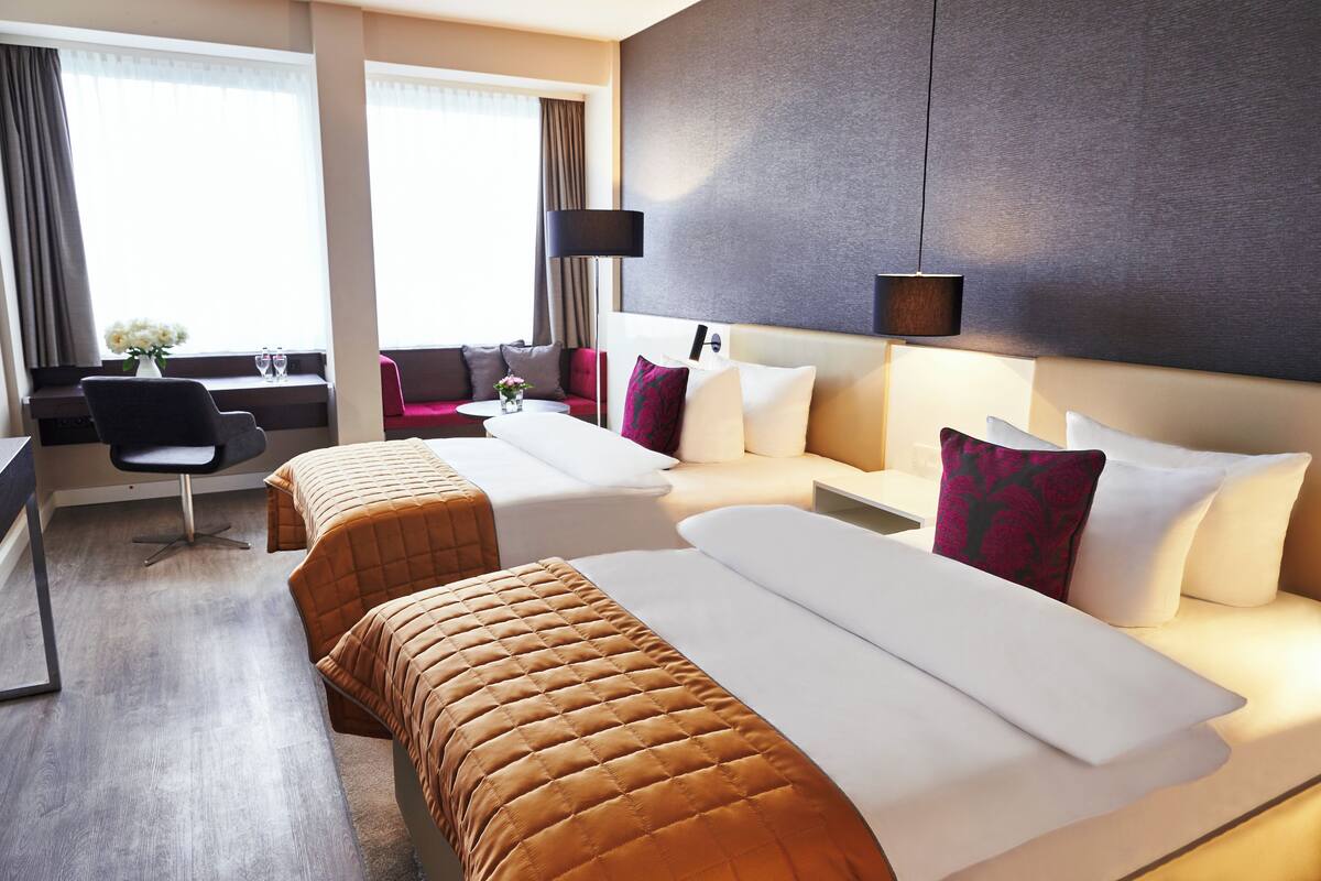Una habitación de hotel con dos camas, un escritorio, una silla y un ventanal grande con cortinas.