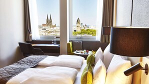A hotel room with a bed, a desk, and a view of a cityscape. - Steigenberger Hotel Köln (Cologne)