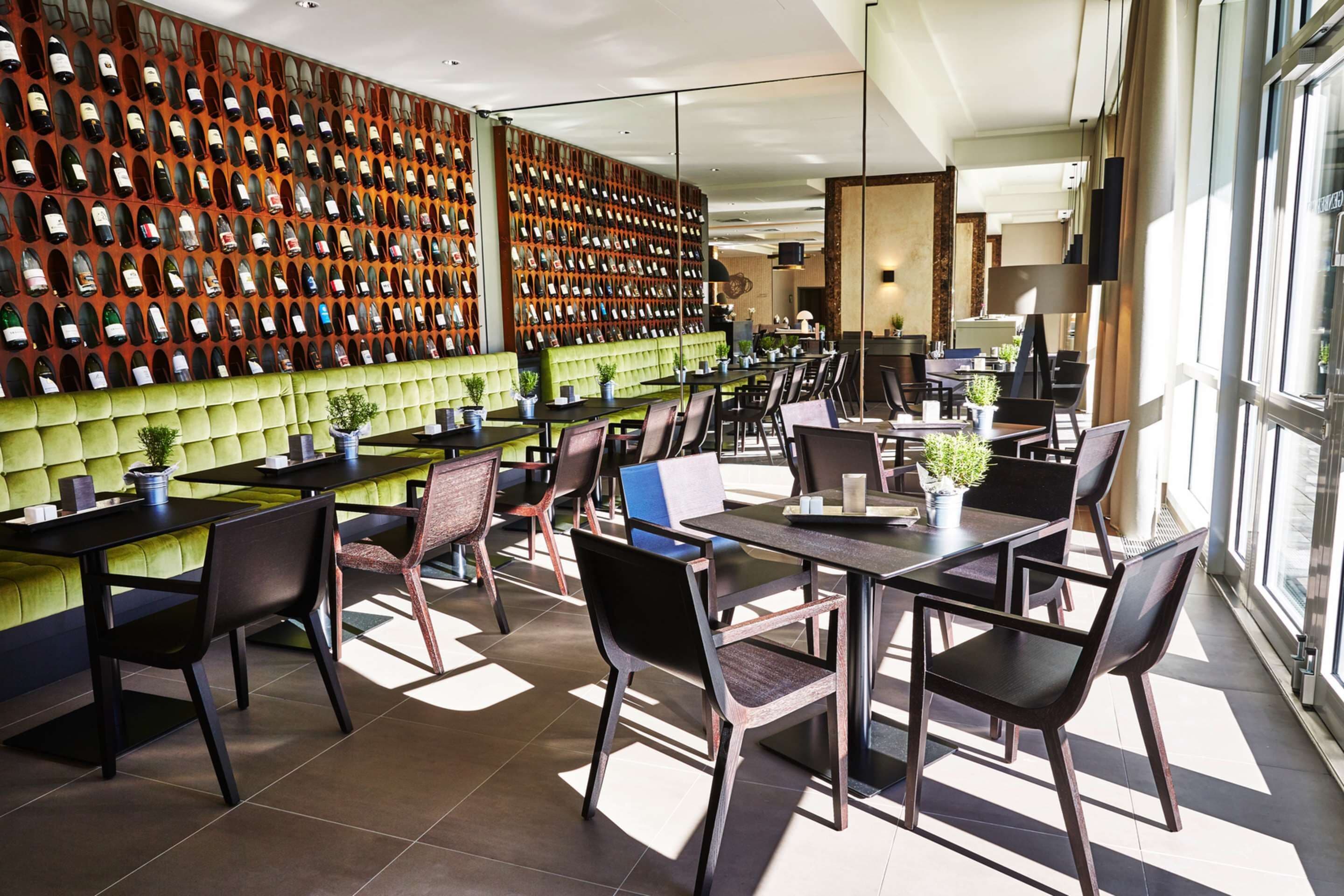 A modern restaurant with a wall of wine bottles, green cushioned seating, and large windows.