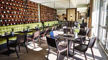 A modern restaurant with a wall of wine bottles, green cushioned seating, and large windows.