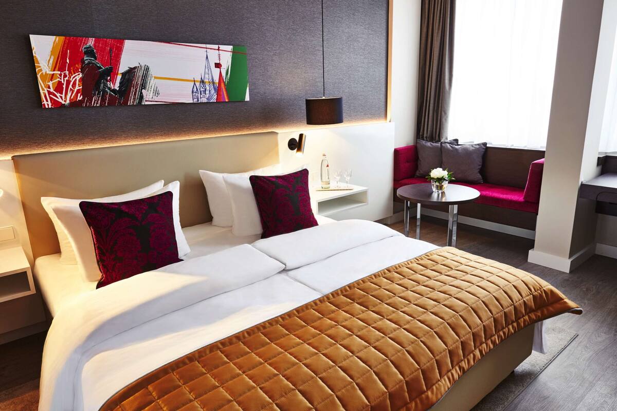 Un cuarto de hotel con una cama grande, un sofá rojo, una mesita con un jarrón de flores y un cuadro en la pared.