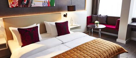 A hotel room with a large bed, a red sofa, a small table with a vase of flowers, and a painting on the wall.