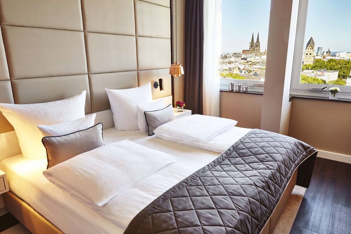 Habitación de hotel con una cama grande, una mesita de noche, una lámpara y vistas a un paisaje urbano.