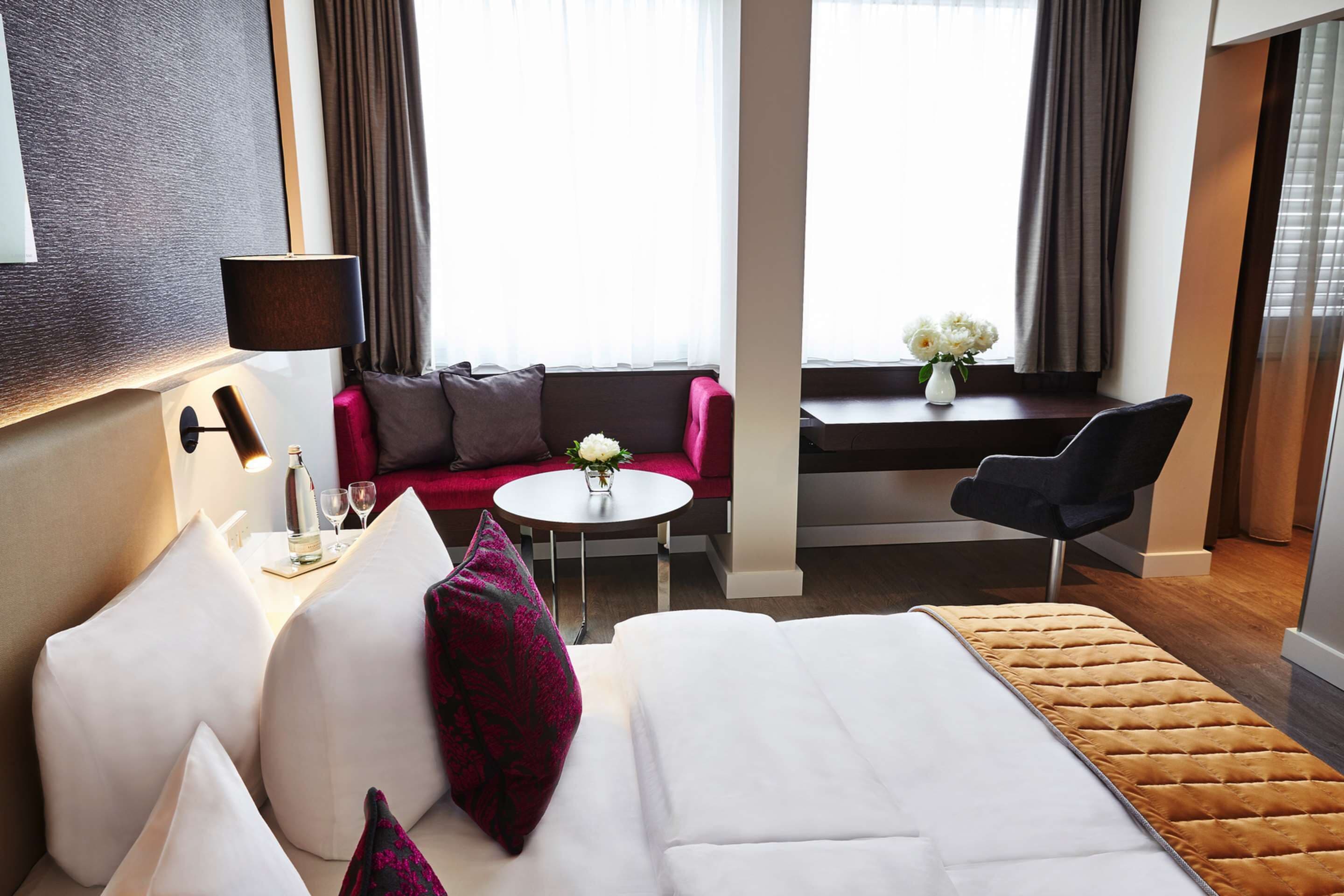 Une chambre d’hôtel avec un lit, un canapé, une petite table sur laquelle est posé un vase de fleurs, une lampe et une chaise.