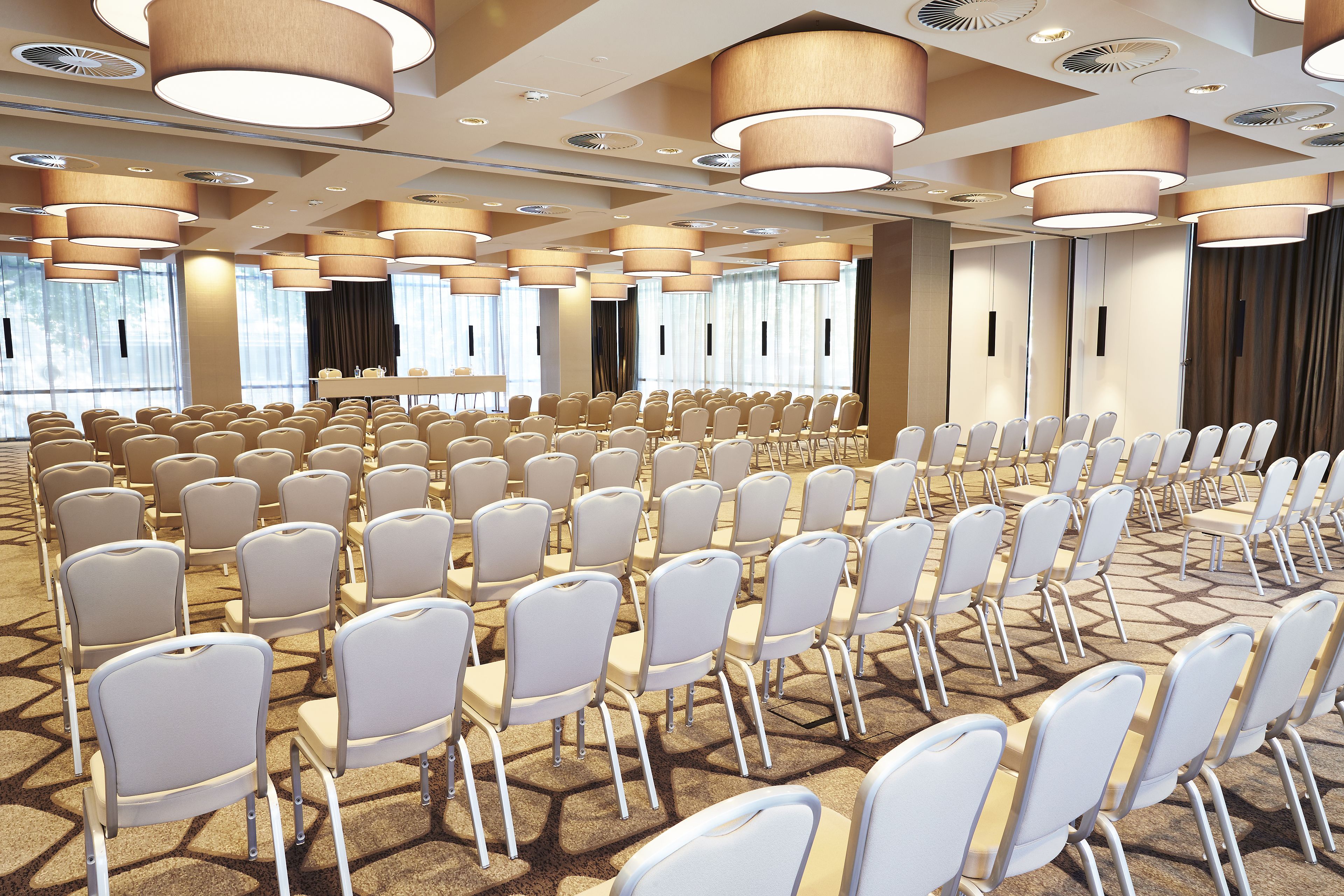 A conference room with rows of white chairs, large ceiling lights, and a carpeted floor.