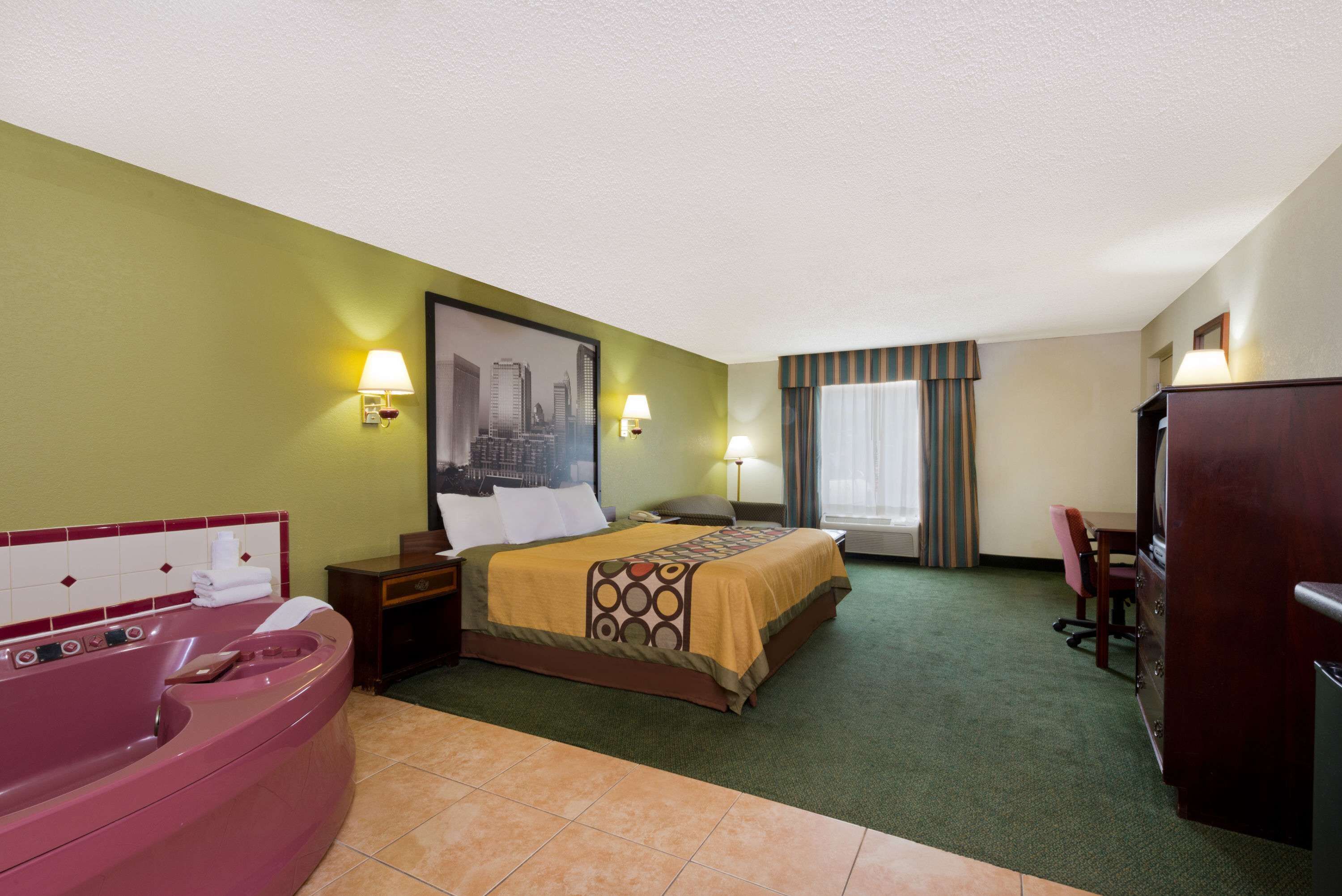 A hotel room with a bed, a large window with curtains, a desk, a chair, a desk lamp, a nightstand, a vanity with a mirror, and a bathtub.