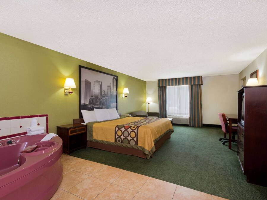 A hotel room with a bed, a large window with curtains, a desk, a chair, a desk lamp, a nightstand, a vanity with a mirror, and a bathtub.