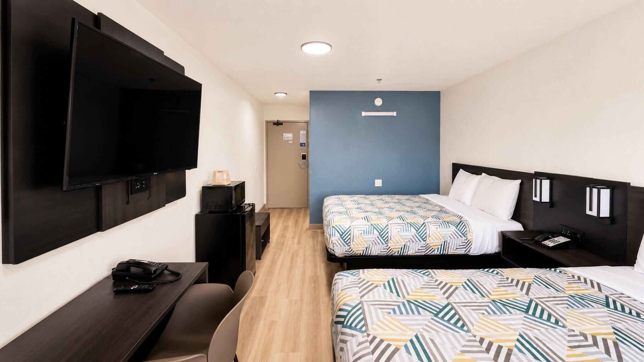 Habitación de hotel con dos camas, un escritorio con teléfono, un televisor de pantalla plana montado en la pared y una pequeña zona de kitchenette.