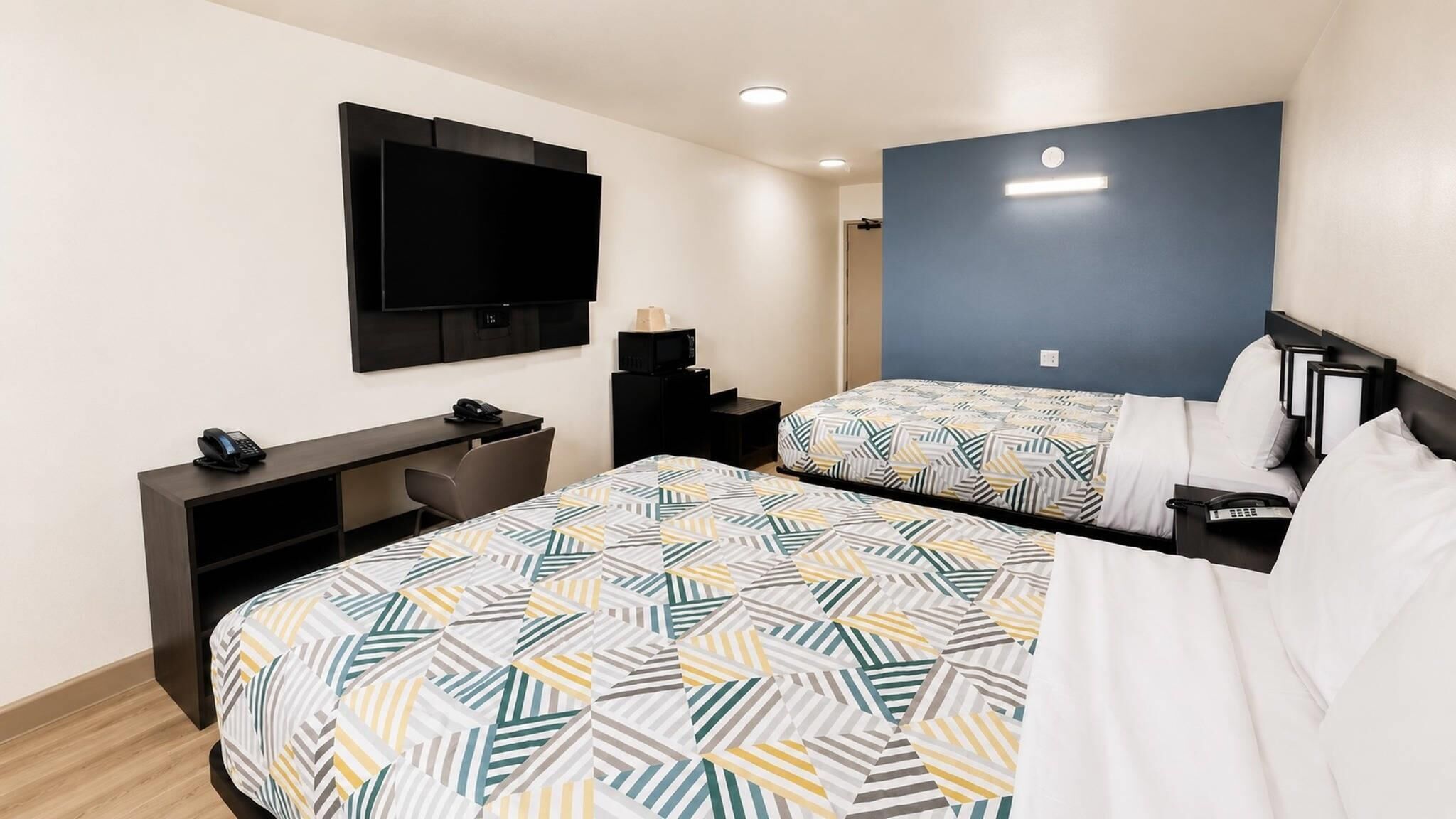 Habitación de hotel con dos camas, un escritorio, un televisor de pantalla plana, una mesita de noche con lámpara y un televisor montado en la pared.