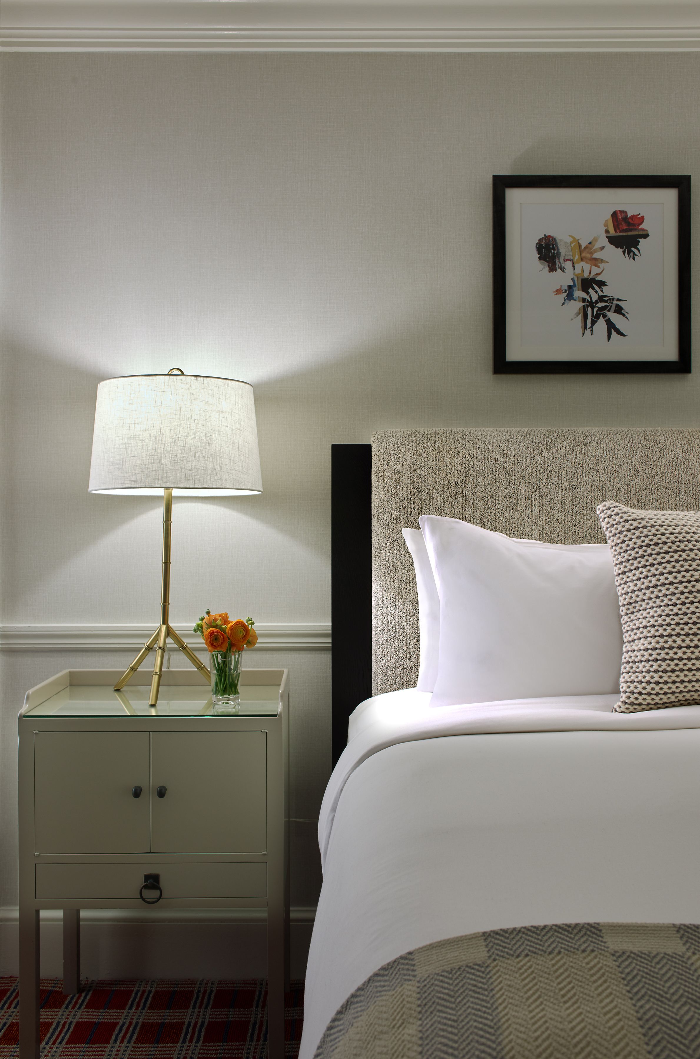 A neatly arranged hotel room with a bedside table, a lamp, a vase with flowers, a bed with pillows, and a framed picture on the wall.