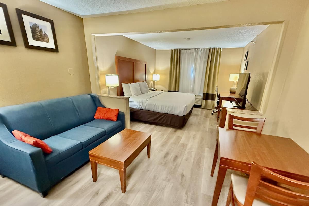 A hotel room with a blue sofa, a wooden coffee table, a bed with white bedding, a wooden desk, and a chair.