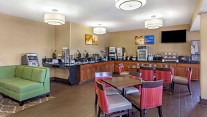 A hotel lobby with a green sofa, a dining area with red and brown chairs, a kitchen area with a coffee machine, and a flat-screen TV on the wall.