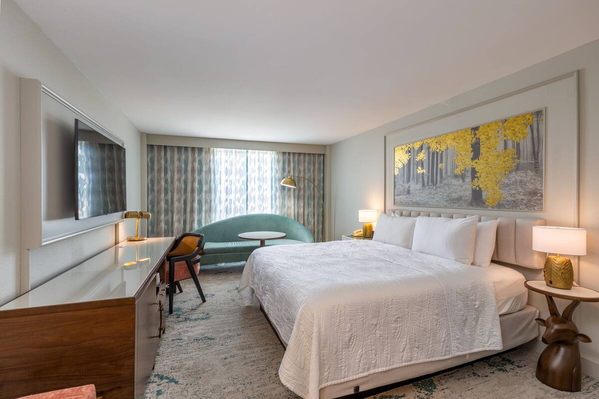 A hotel room with a large bed, a desk, a chair, a small table, and a painting on the wall.