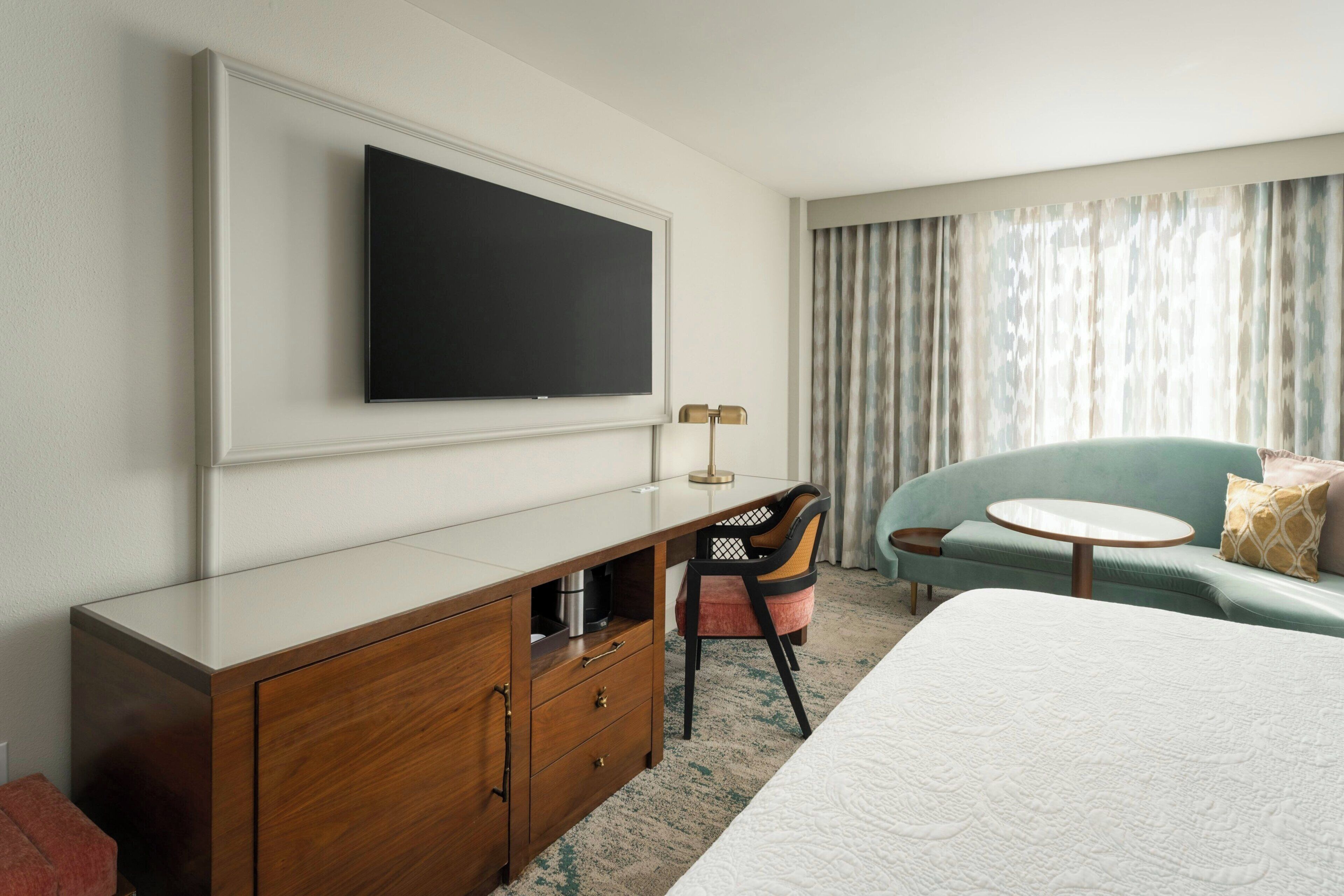 A hotel room with a bed, a desk with a flat-screen TV, a chair, a sofa, and a round table.