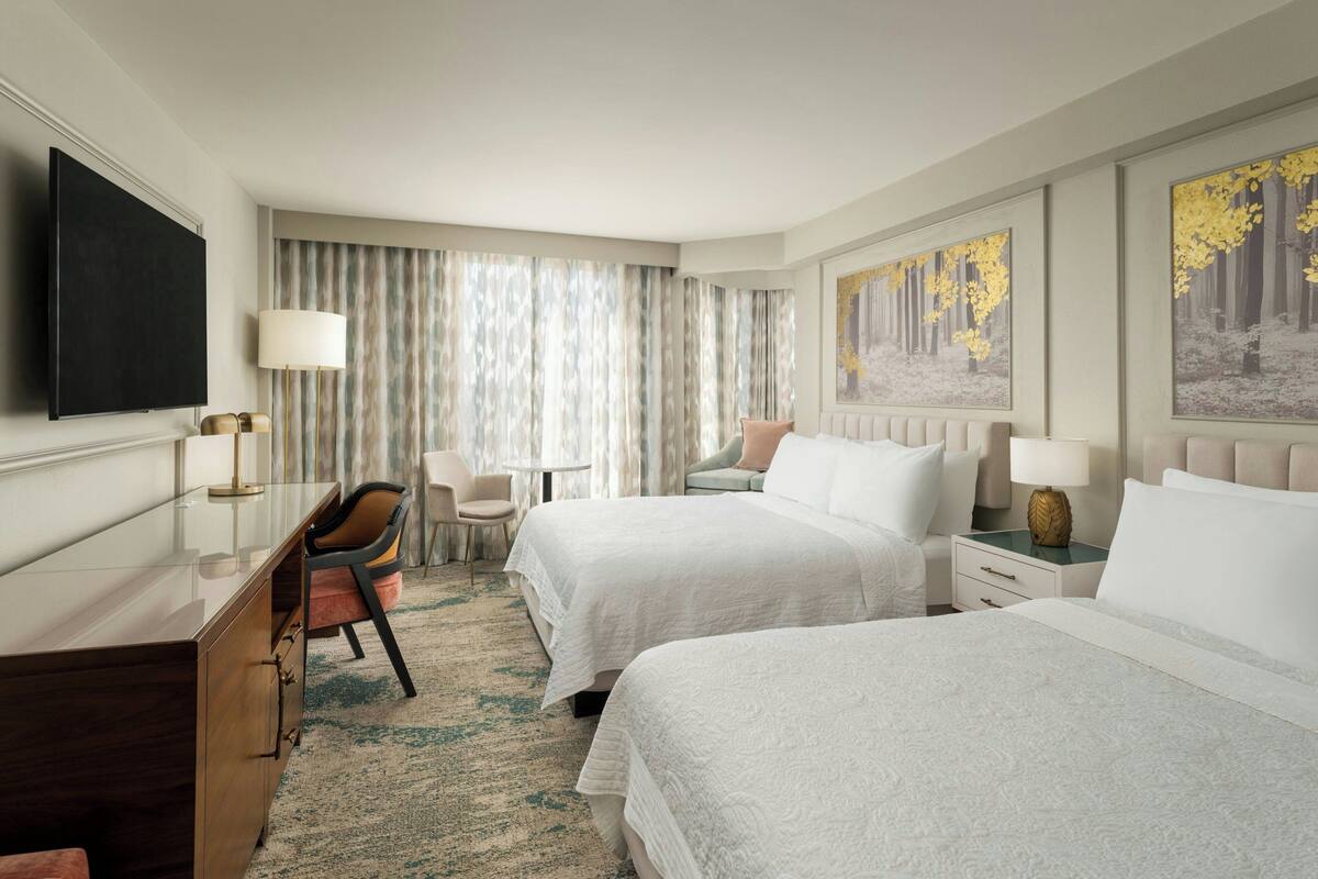 A hotel room with two beds, a desk, a chair, a TV, and artwork on the walls.