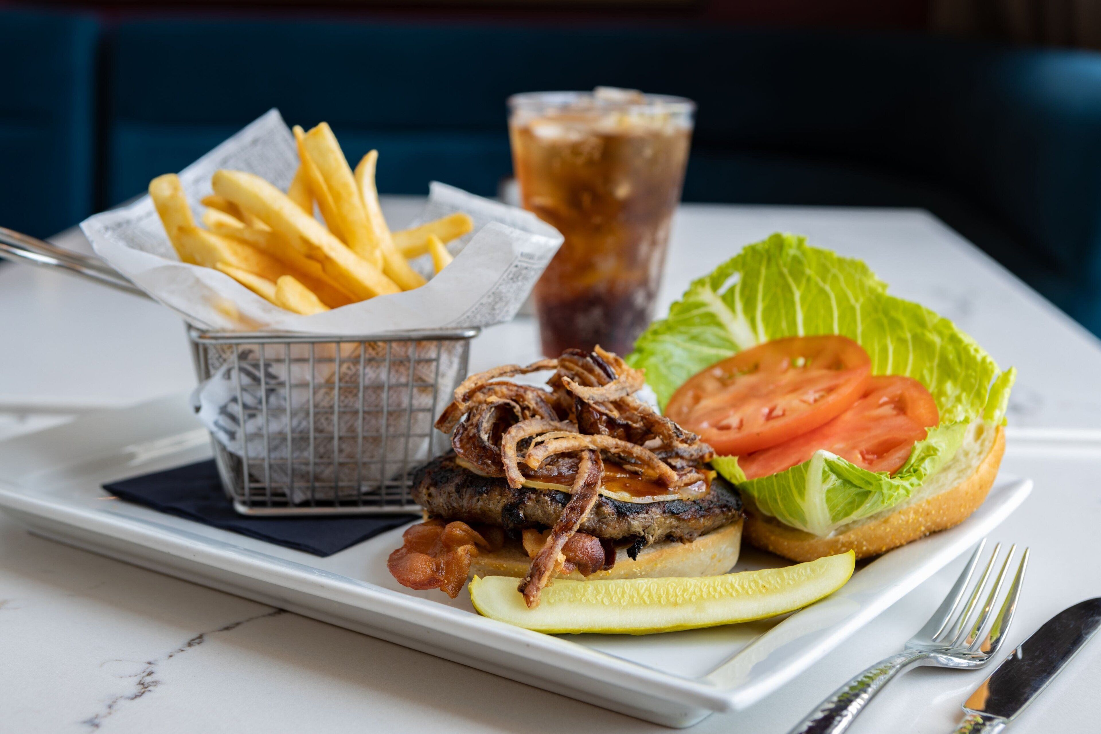 A meal consisting of a burger with onions and bacon, a side of fries, a slice of tomato, and a pickle on a white plate.