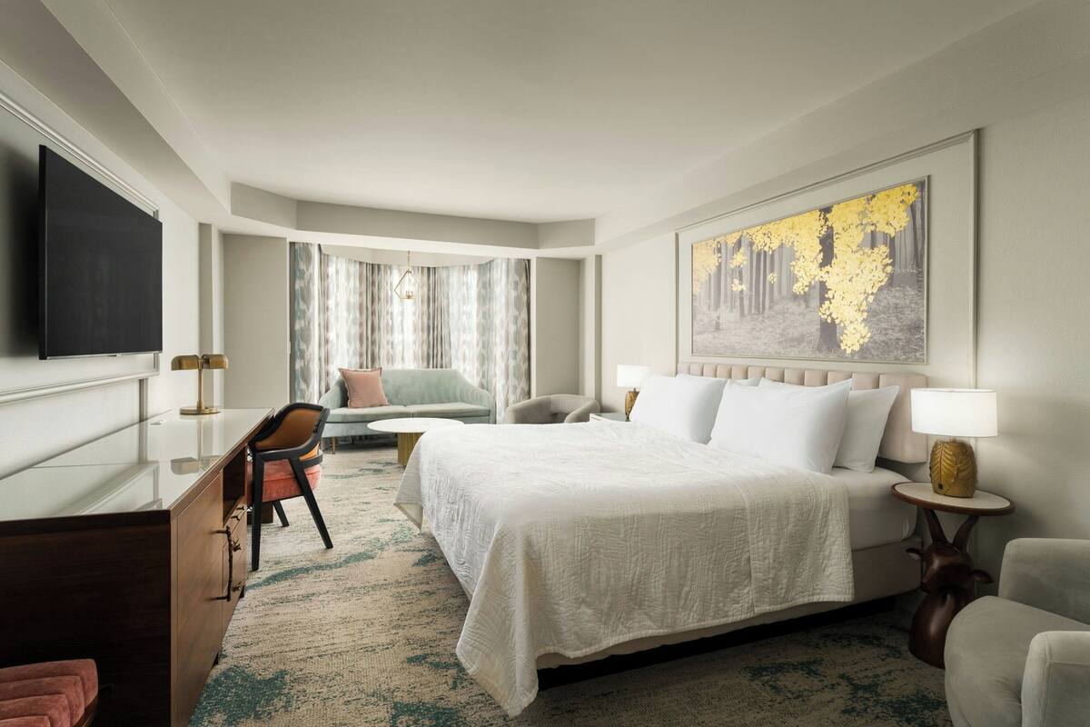 A hotel room with a bed, a desk, a chair, a TV, and a painting on the wall.