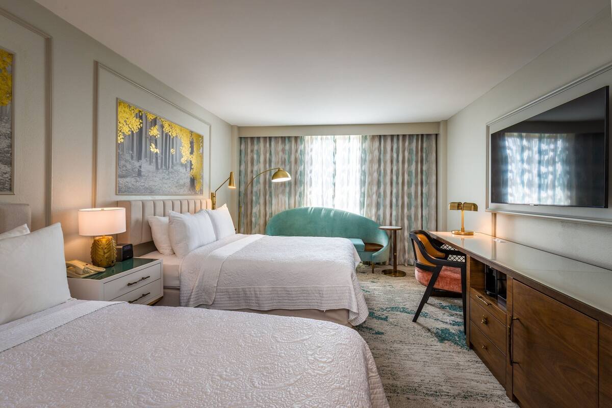 A hotel room with two beds, a desk, a chair, a TV, and a painting on the wall.